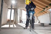 Seseorang yang mengenakan perlengkapan keselamatan menggunakan palu pembongkaran untuk menghancurkan lantai beton.