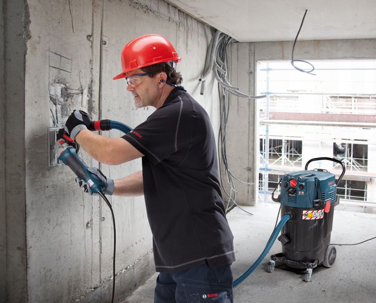 Seseorang yang mengenakan peralatan keselamatan sedang menggiling dinding beton, sementara alat penyedot debu beroperasi di dekatnya.