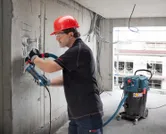 Seseorang yang mengenakan peralatan keselamatan sedang menggiling dinding beton, sementara alat penyedot debu beroperasi di dekatnya.