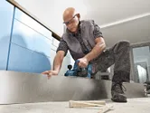 Seseorang yang mengenakan perlengkapan keselamatan sedang meratakan papan kayu menggunakan planer tanpa kabel.