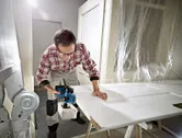 Seseorang yang mengenakan perlengkapan keselamatan menggunakan planer tanpa kabel untuk menghaluskan pintu putih.