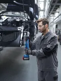 Seseorang yang mengenakan peralatan keselamatan mengencangkan baut pada rangka mobil menggunakan alat listrik tanpa kabel.