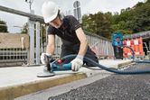 Seseorang yang mengenakan peralatan keselamatan menggiling beton di trotoar dengan penggiling yang terhubung.