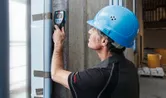 Seseorang yang mengenakan peralatan keselamatan memindai dinding beton dengan detektor pemindai dinding.