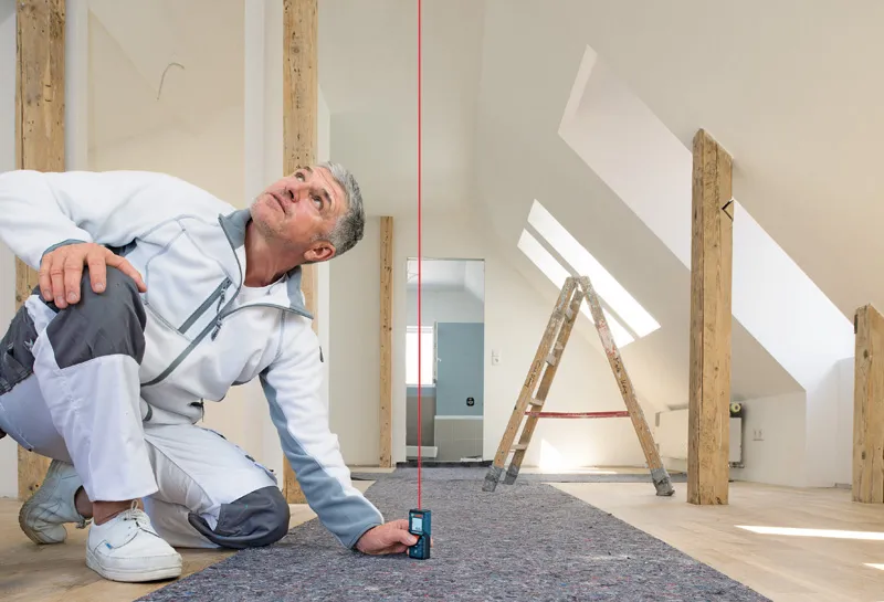 Seseorang menggunakan pengukur laser untuk memeriksa keselarasan vertikal di ruangan loteng yang terang.