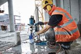 Seseorang yang mengenakan peralatan keselamatan mengoperasikan bor berlian di lantai beton basah.