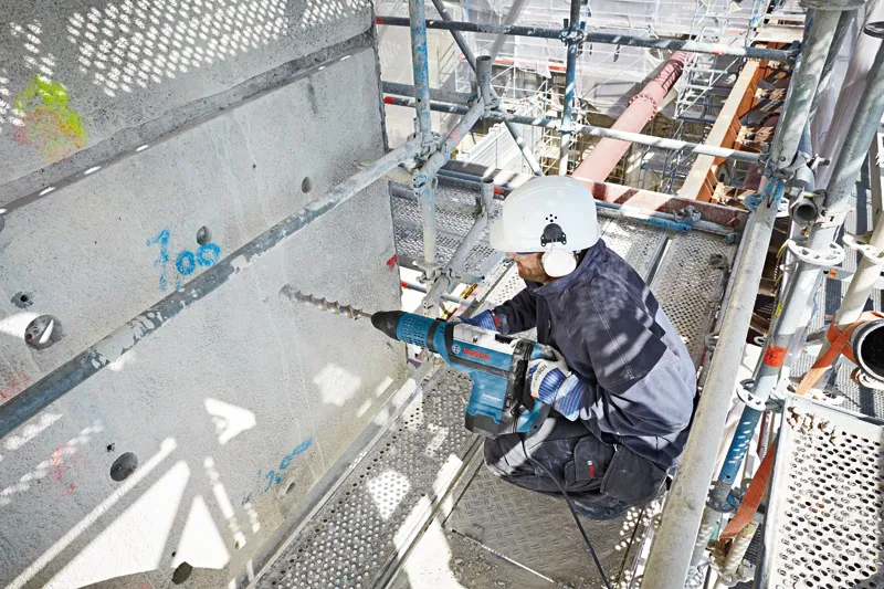 Seseorang yang mengenakan peralatan keselamatan mengebor beton dengan palu putar besar.