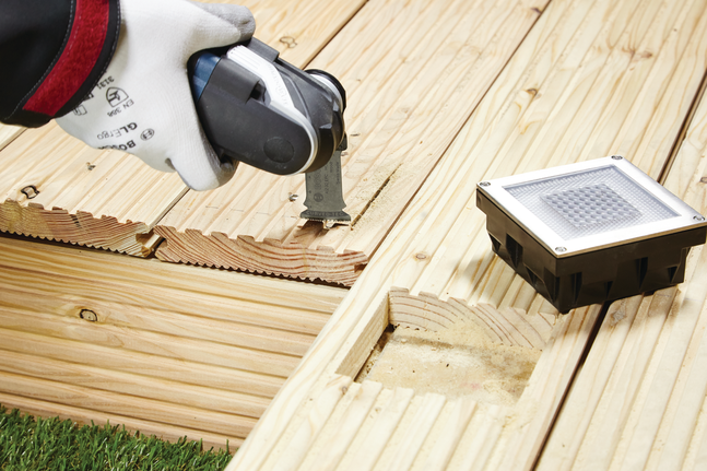 Pekerja yang mengenakan perlengkapan keselamatan memotong alur pada dek kayu menggunakan alat serbaguna.