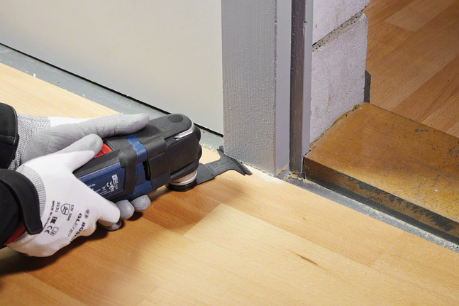 Orang yang mengenakan peralatan keselamatan memangkas lantai kayu di samping kusen pintu menggunakan perkakas listrik.