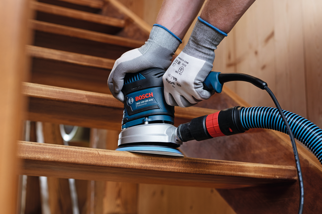 Seorang pekerja yang mengenakan perlengkapan keselamatan mengampelas tapak tangga kayu menggunakan mesin pengamplas listrik yang terhubung.