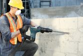 Seseorang yang mengenakan perlengkapan keselamatan menggunakan palu putar untuk mengebor dinding beton.