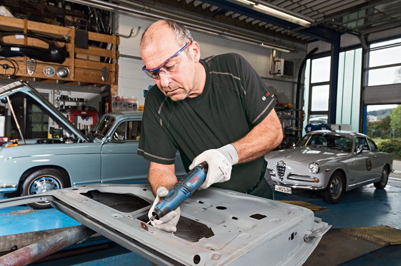 Seseorang yang mengenakan perlengkapan keselamatan menggunakan alat putar tanpa kabel untuk menggiling logam di garasi.