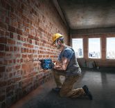 Seseorang yang mengenakan peralatan keselamatan mengebor dinding bata di dalam gedung yang sedang dibangun.