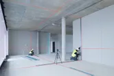 Dua orang yang mengenakan peralatan keselamatan menyelaraskan drywall menggunakan alat perata laser di sebuah gedung.