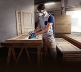 Seseorang yang mengenakan perlengkapan keselamatan mengampelas pintu kayu dengan sander orbital di bengkel.