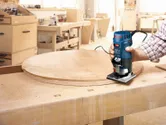 Seseorang memandu router telapak tangan di sepanjang tepi papan kayu bundar pada meja kerja.