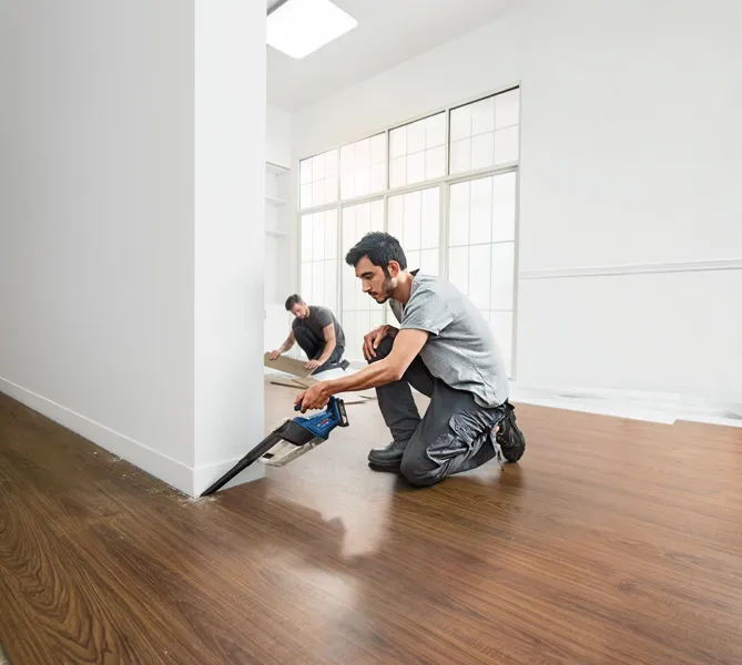 Seseorang memotong lantai di dekat dinding dengan gergaji tanpa kabel sementara yang lain memasang panel lantai.