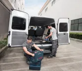 Dua pekerja di dekat mobil van terbuka mengatur peralatan dan menggunakan penyedot debu tanpa kabel.