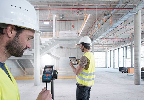 GLM 150 C Dua orang yang mengenakan peralatan keselamatan menggunakan pengukur laser dan tablet di dalam lokasi konstruksi besar.