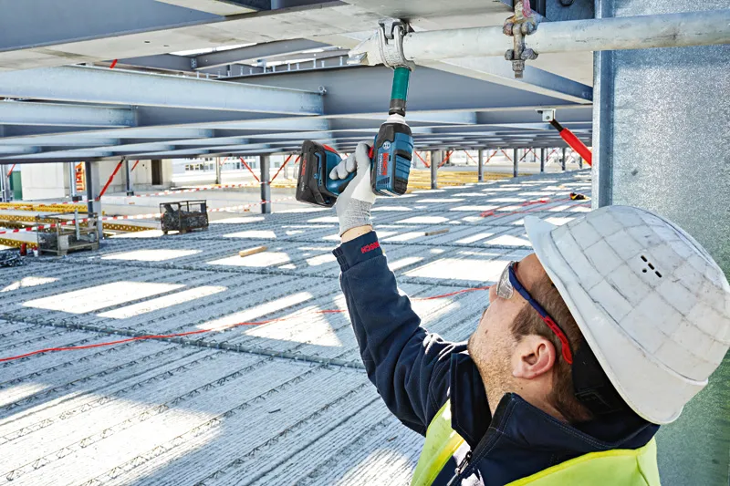 Seseorang yang mengenakan perlengkapan keselamatan menggunakan kunci pas benturan tanpa kabel pada balok logam.