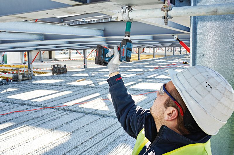 Seseorang yang mengenakan perlengkapan keselamatan menggunakan kunci pas benturan tanpa kabel pada balok logam.