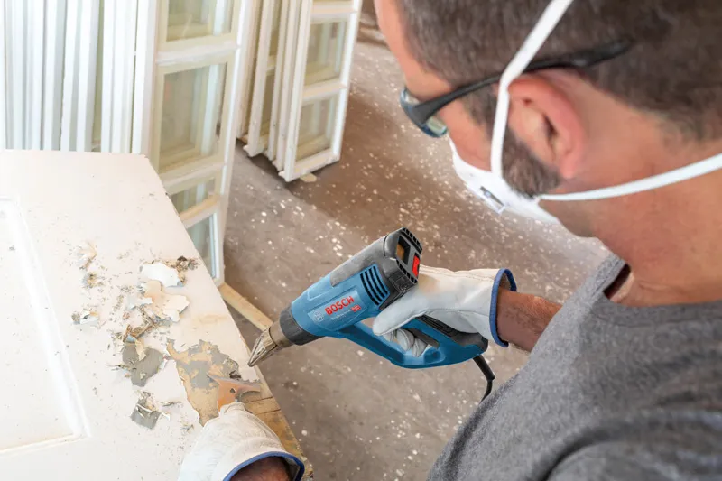 Orang yang mengenakan perlengkapan keselamatan menggunakan senapan panas untuk mengelupas cat dari pintu.