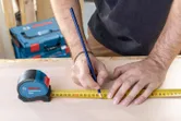 Orang menandai ukuran pada kayu dengan pensil menggunakan pita pengukur.