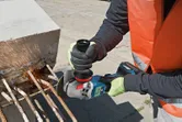 Seseorang yang mengenakan perlengkapan keselamatan sedang menggiling tulangan beton yang memanjang dari balok beton.