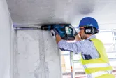 Seseorang yang mengenakan peralatan keselamatan mengebor langit-langit beton dengan palu putar.