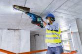 Seseorang yang mengenakan peralatan keselamatan mengebor langit-langit beton dengan palu putar tanpa kabel.