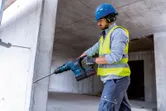Seseorang yang mengenakan perlengkapan keselamatan menggunakan palu putar tanpa kabel untuk mengebor dinding beton.