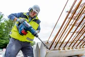 Seseorang yang mengenakan peralatan keselamatan mengebor beton dengan palu putar besar tanpa kabel.