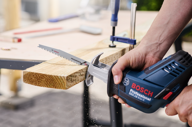 Seseorang menggunakan gergaji bolak-balik untuk memotong papan kayu di atas meja kerja.