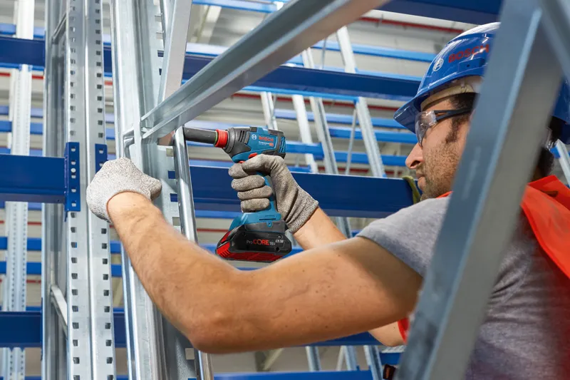 Seseorang yang mengenakan peralatan keselamatan menggunakan bor tanpa kabel pada rangka logam.