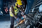 Seseorang yang mengenakan perlengkapan keselamatan menggunakan penggiling sudut tanpa kabel untuk memotong tulangan beton.