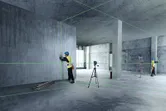 Dua pekerja yang mengenakan peralatan keselamatan menyelaraskan dinding bagian dalam menggunakan alat perataan laser.
