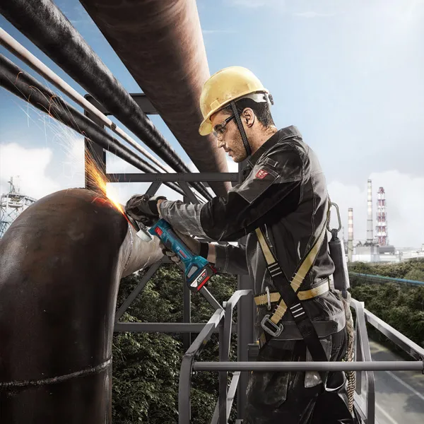 Seseorang yang mengenakan peralatan keselamatan menggiling pipa logam besar dengan penggiling sudut.