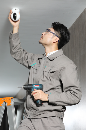 Seseorang yang mengenakan peralatan keselamatan sedang memasang kamera keamanan dengan menggunakan bor.