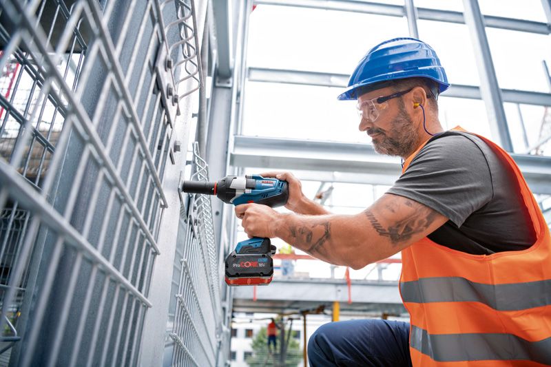 Seseorang yang mengenakan perlengkapan keselamatan menggunakan kunci pas benturan tanpa kabel pada pagar logam.