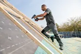 Seseorang yang mengenakan peralatan keselamatan menggunakan bor tanpa kabel untuk mengencangkan kayu pada atap miring.