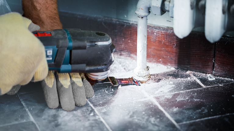 Seseorang yang mengenakan perlengkapan keselamatan menggunakan alat listrik untuk memotong lantai di sekitar pipa.