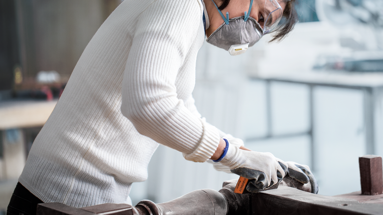 Seseorang yang mengenakan alat pengaman mengampelas permukaan kayu dengan alat tangan.