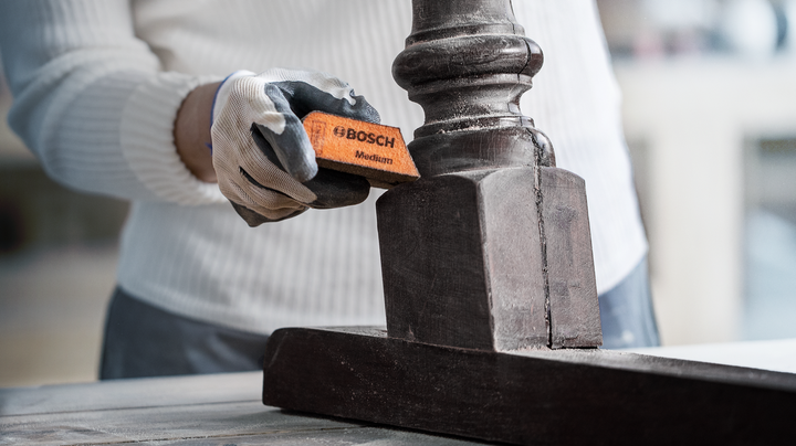 Object #200360618: 2793255_web_002 Seorang pekerja yang mengenakan peralatan keselamatan sedang mengampelas kaki furnitur kayu.