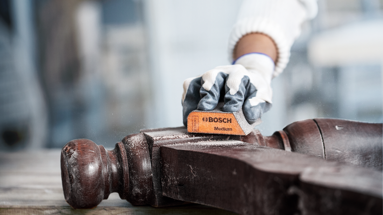 Seseorang yang mengenakan peralatan keselamatan sedang mengampelas sebuah perabot kayu.