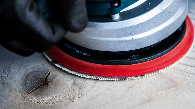 Seseorang yang mengenakan peralatan keselamatan mengampelas permukaan kayu dengan mesin pengampelas listrik.