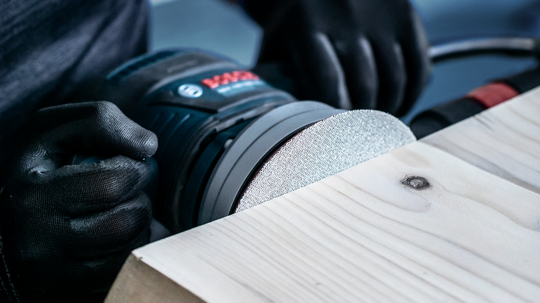 Seseorang yang mengenakan alat pengaman mengamplas papan kayu dengan mesin amplas listrik.