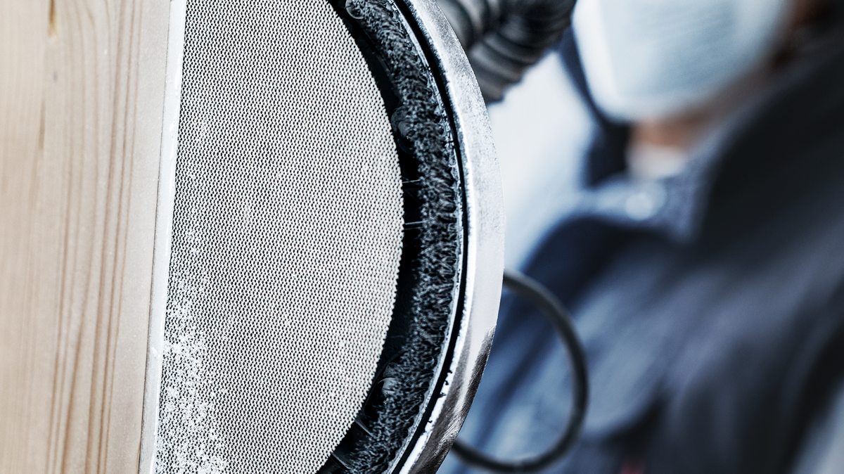 Seseorang yang mengenakan peralatan keselamatan mengamplas kayu dengan mesin amplas bundar bertenaga listrik.