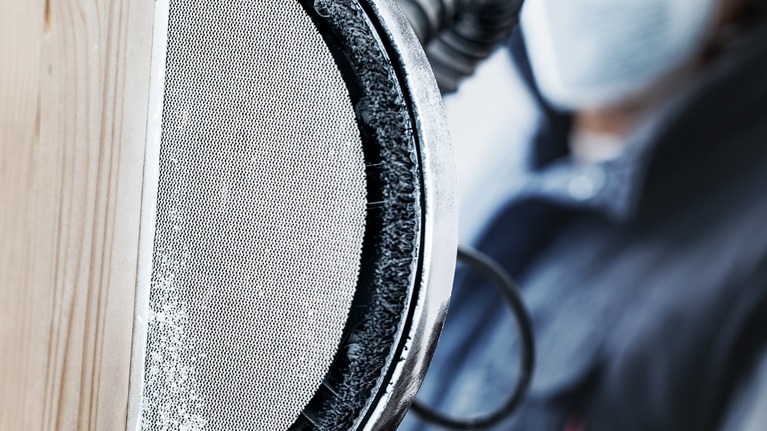 Seseorang yang mengenakan peralatan keselamatan mengamplas kayu dengan mesin amplas bundar bertenaga listrik.