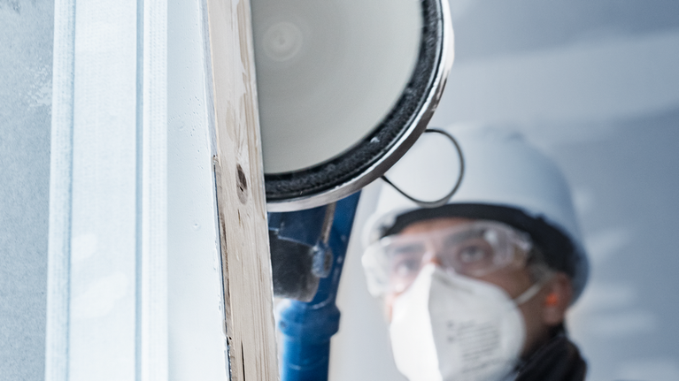 Seseorang yang mengenakan peralatan keselamatan menggunakan perkakas listrik untuk memotong permukaan kayu.