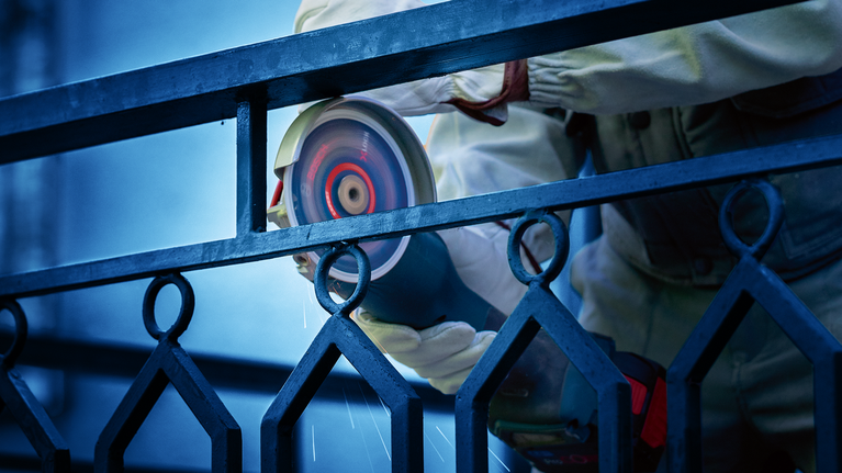 Seseorang yang mengenakan peralatan keselamatan sedang menggerinda pagar logam menggunakan alat listrik.